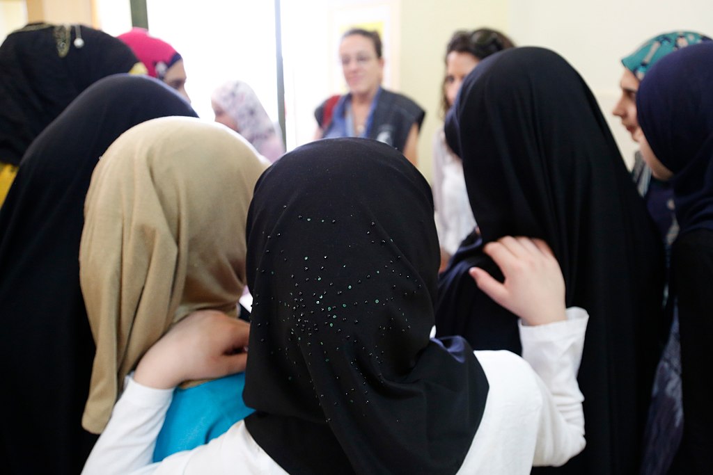 A group of young women, wearing hijabs of various colors, stand in a circle during a discussion at a community center. 